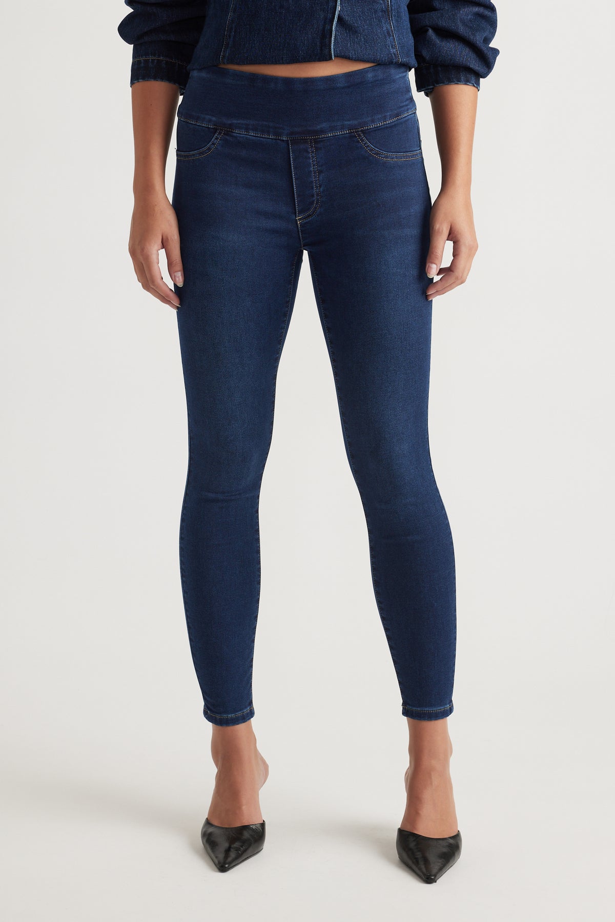 Front view of woman wearing dark indigo skinny crop jeans paired with black pointed-toe heels and denim jacket