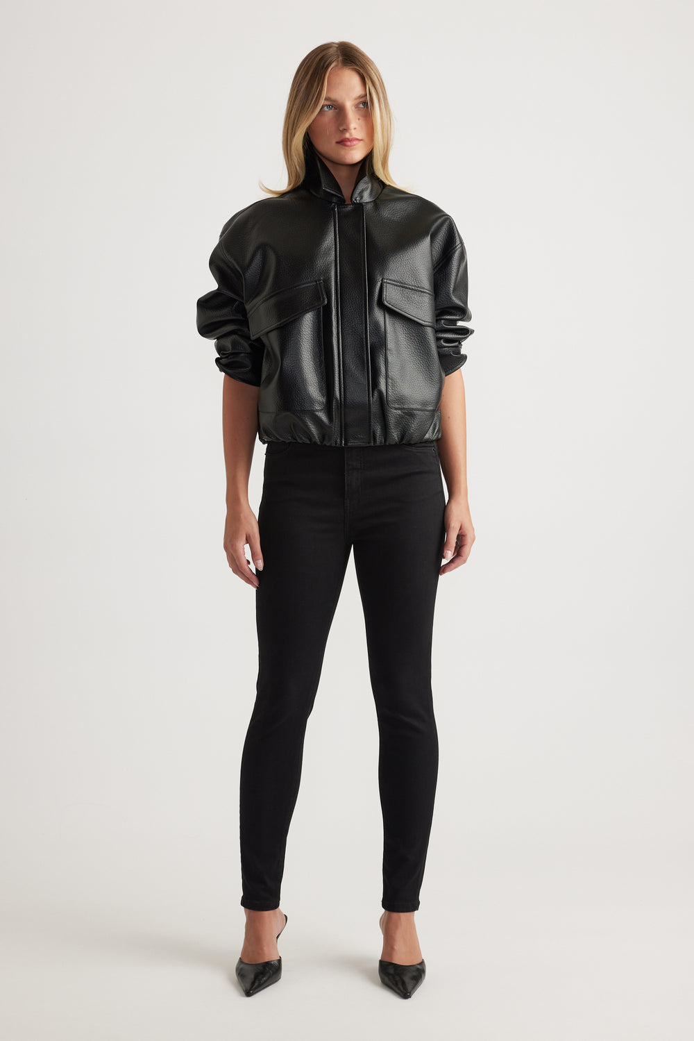 Woman wearing a black faux leather bomber jacket with oversized pockets paired with slim black pants and pointed heels