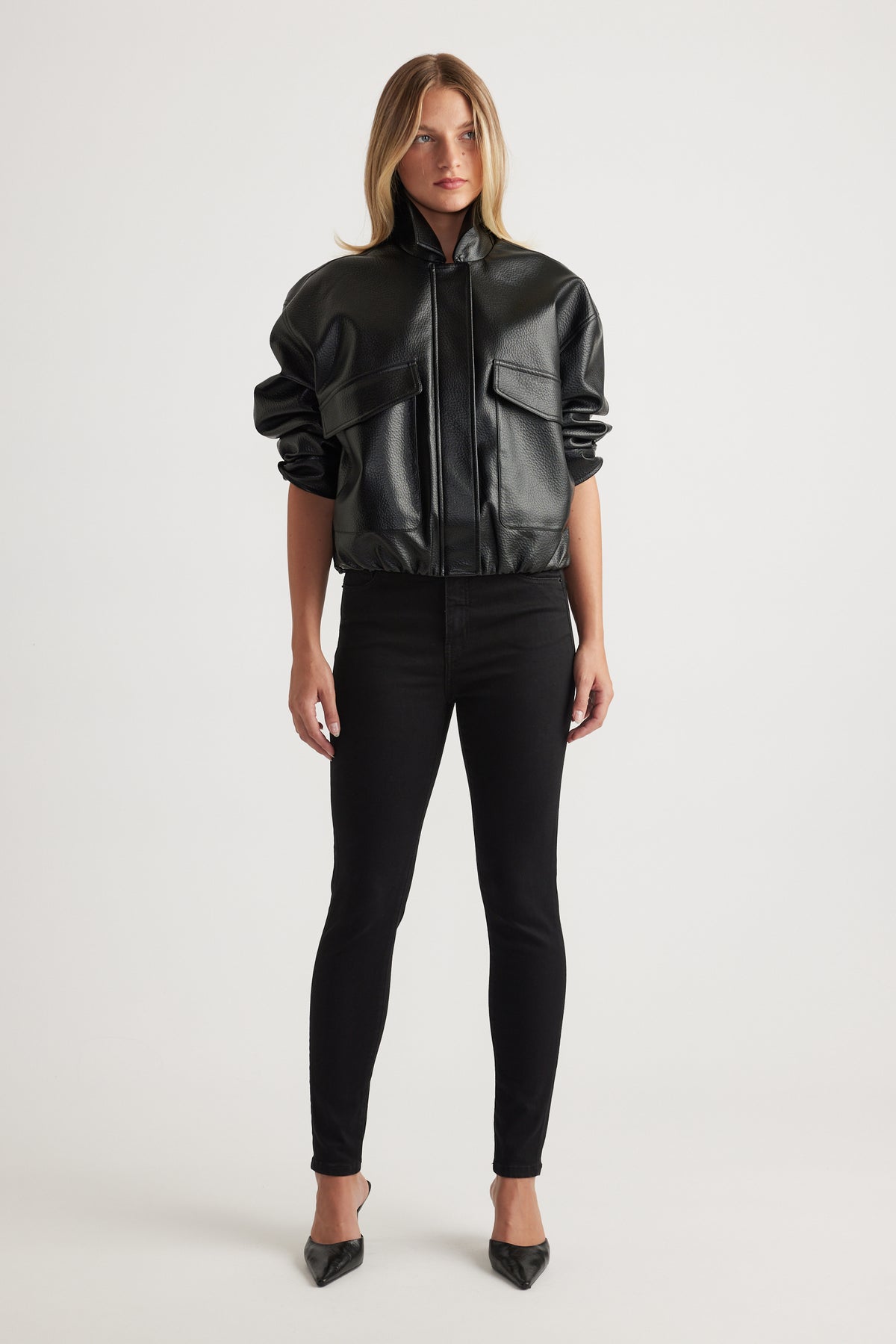 Woman wearing a black faux leather bomber jacket with oversized pockets paired with slim black pants and pointed heels