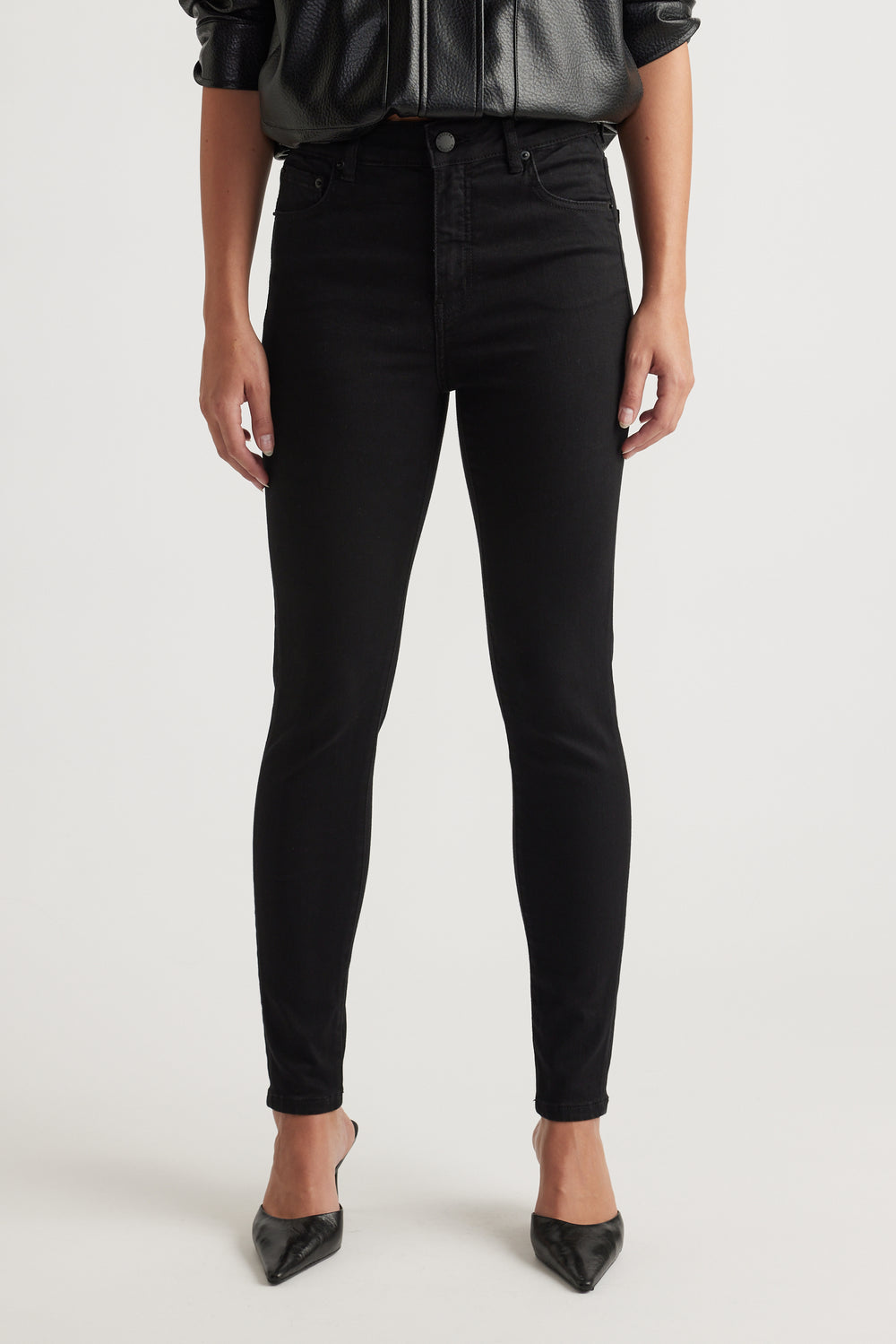 Woman wearing slim black jeans paired with pointed black heels and a black leather-look blouse, front view.