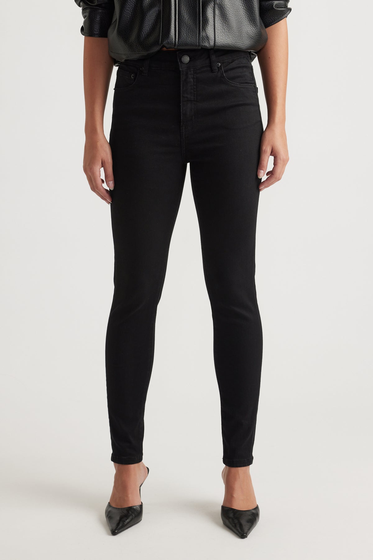 Woman wearing slim black jeans paired with pointed black heels and a black leather-look blouse, front view.