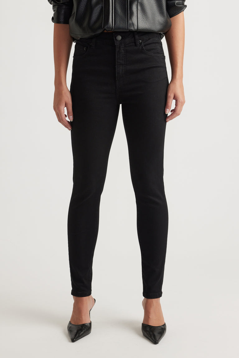 Woman wearing slim black jeans paired with pointed black heels and a black leather-look blouse, front view.