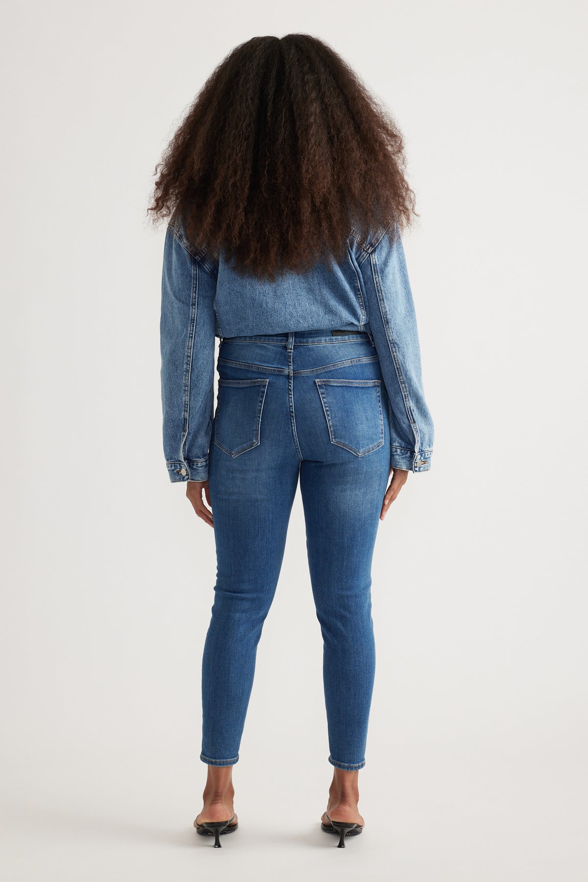 Back view of woman wearing high rise blue skinny jeans with ankle-length legs and a denim jacket