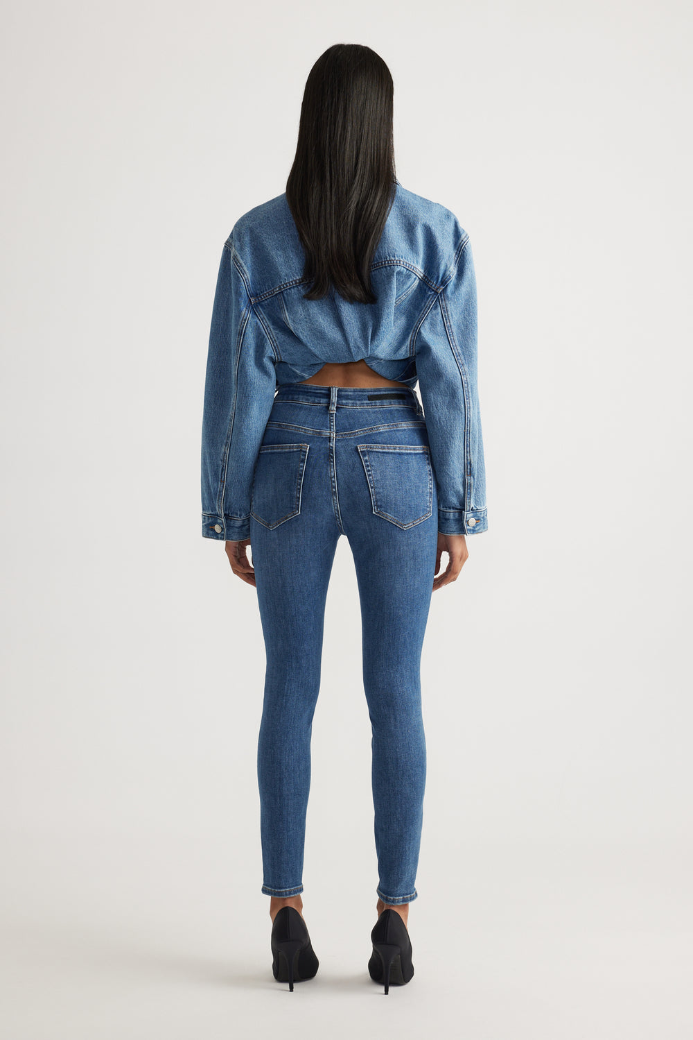 Rear view of model wearing Evie High Rise Skinny Jean in mid blue denim styled with cropped denim jacket by Decjuba Australian brand