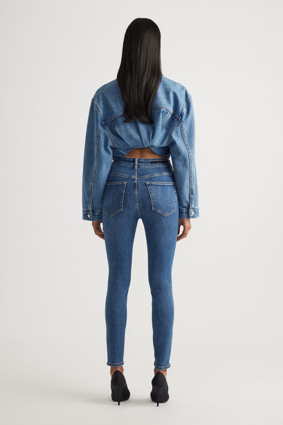 Rear view of model wearing Evie High Rise Skinny Jean in mid blue denim styled with cropped denim jacket by Decjuba Australian brand