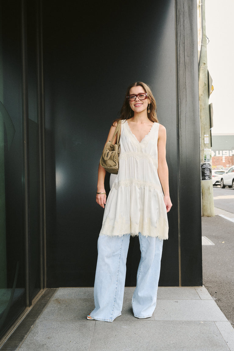 A woman stands on a sidewalk wearing a sleeveless cream lace tiered tunic top, light blue wide-leg jeans, and carrying a tan shoulder bag.