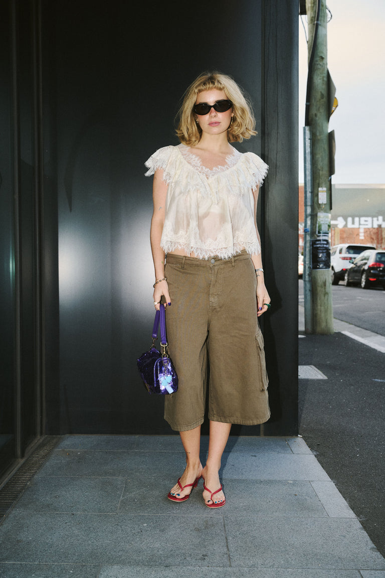 A woman stands on a city sidewalk wearing a sheer white ruffled sleeveless top, olive green cropped cargo pants, red flip-flop sandals, and holding a purple handbag.
