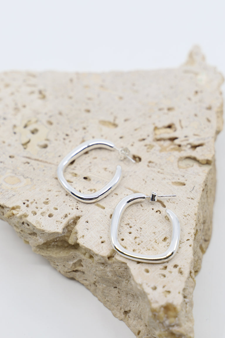 Pair of polished irregular-shaped silver hoop earrings resting on textured natural stone surface.