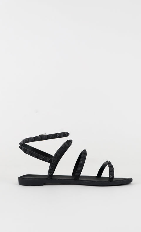 Black flat sandal with multiple studded straps and ankle wrap, featuring a minimalist and edgy summer design.