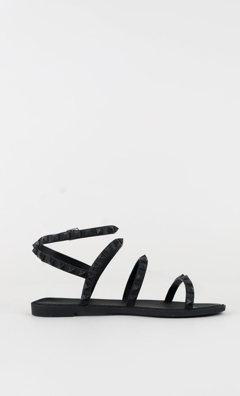 Black flat sandal with multiple studded straps and ankle wrap, featuring a minimalist and edgy summer design.