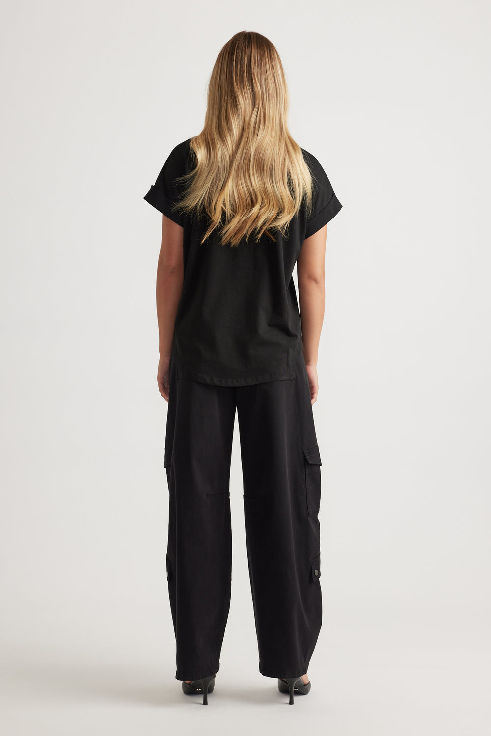 Woman shown from behind wearing relaxed black tee with rolled sleeves paired with wide-leg black cargo pants and heels