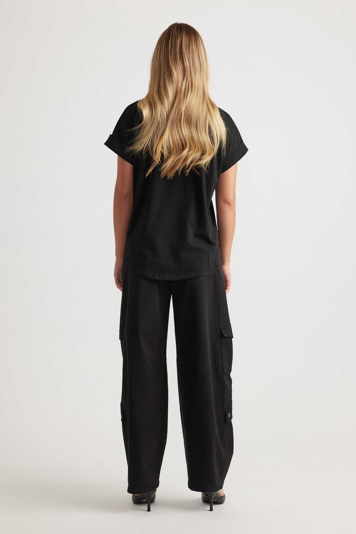 Woman shown from behind wearing relaxed black tee with rolled sleeves paired with wide-leg black cargo pants and heels