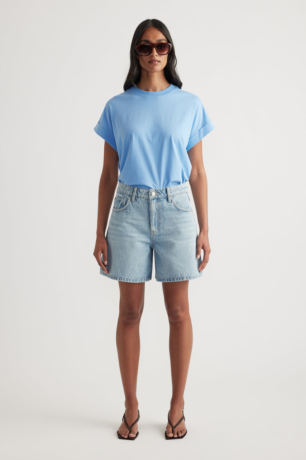 Woman wearing a light blue crew neck tee with denim shorts, sunglasses and strappy sandals
