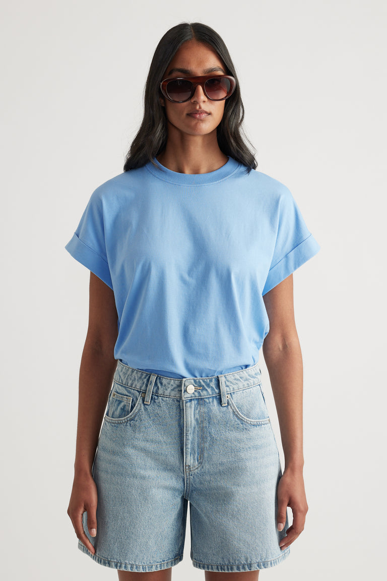 Model wearing a light blue crew neck tee with rolled cuffs, denim shorts and sunglasses