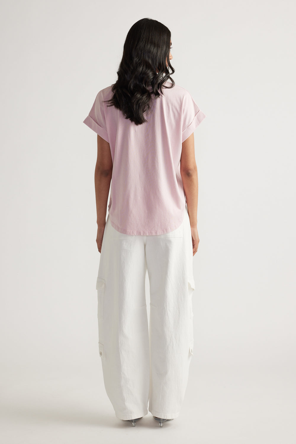 Back view of woman wearing a pale pink crew neck tee with rolled cuffs and relaxed white cargo pants