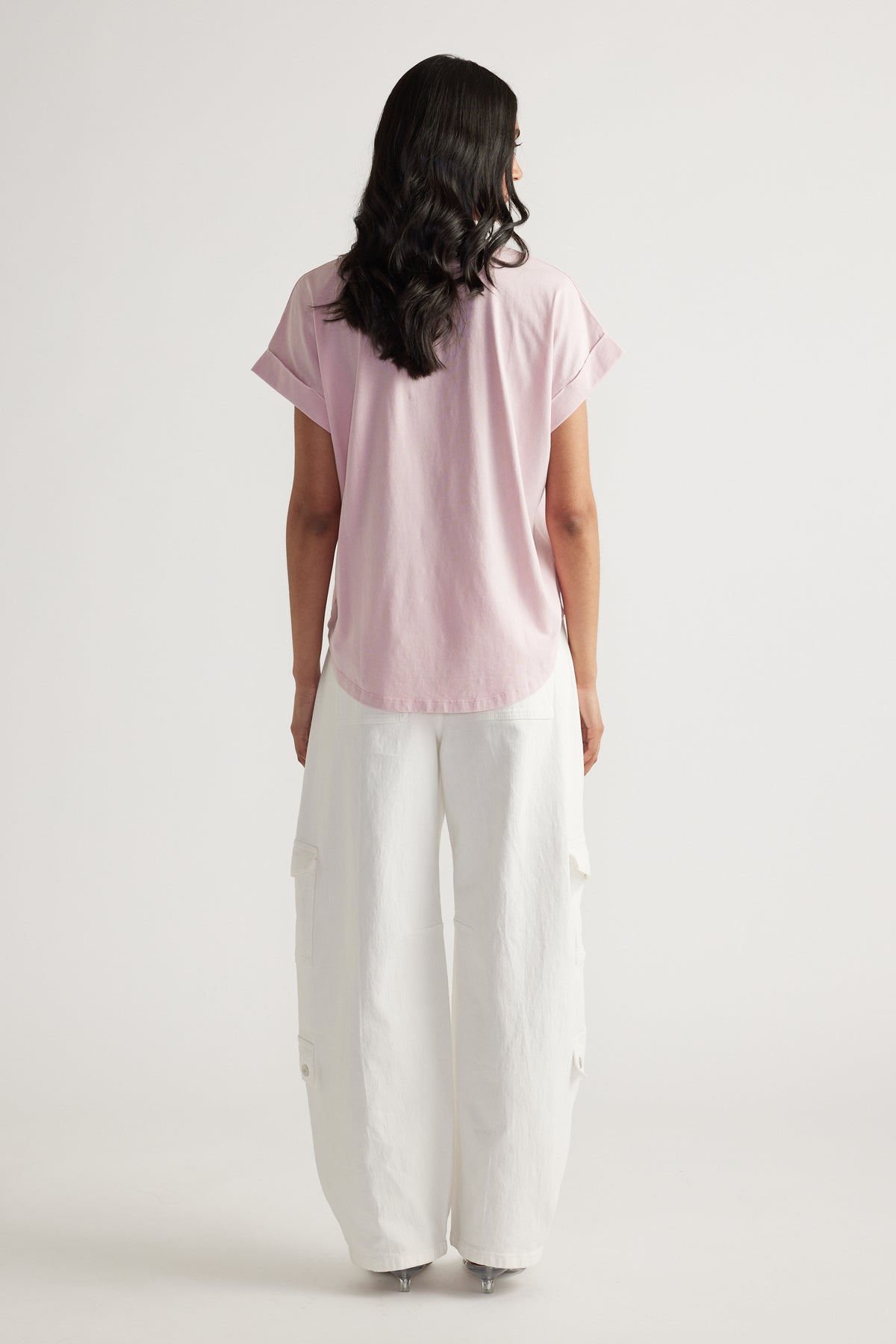 Back view of woman wearing a pale pink crew neck tee with rolled cuffs and relaxed white cargo pants