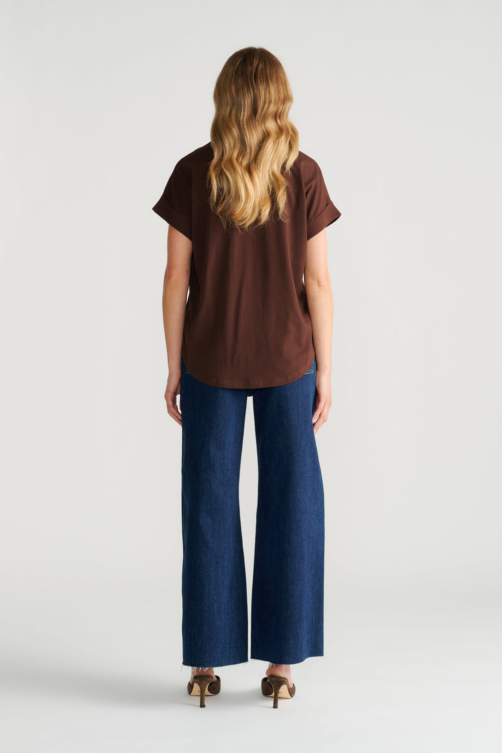 Woman wearing a relaxed fit brown crew neck tee paired with wide-leg dark blue denim jeans and heeled sandals, back view.