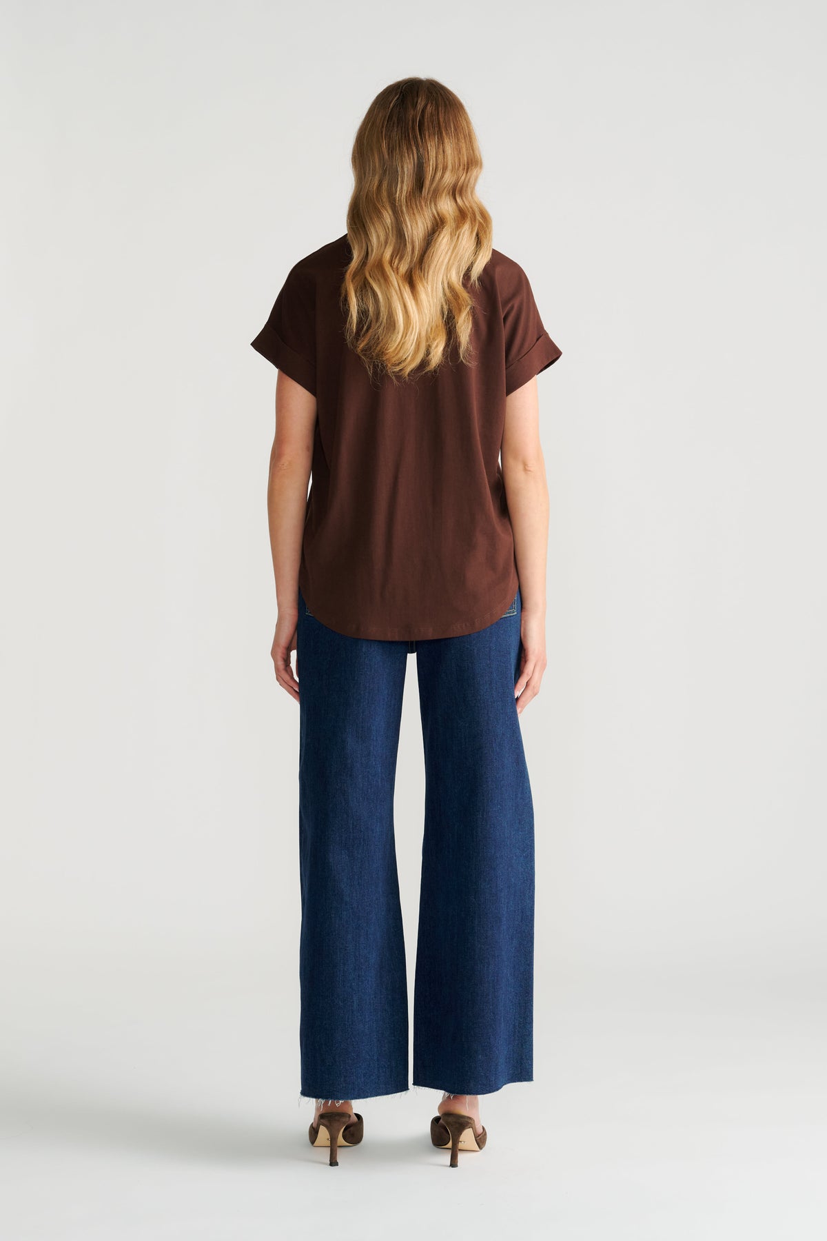 Woman wearing a relaxed fit brown crew neck tee paired with wide-leg dark blue denim jeans and heeled sandals, back view.