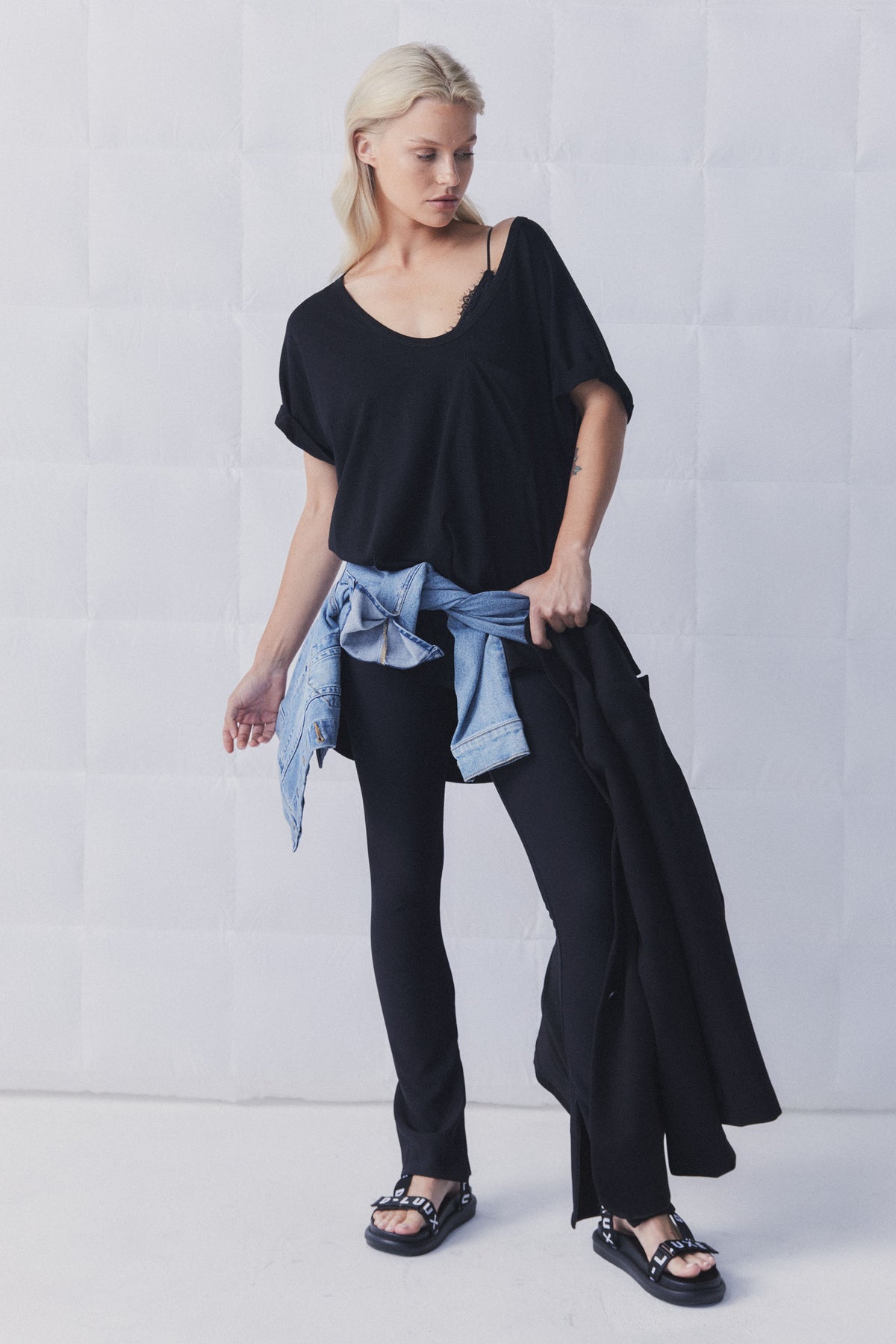 Woman wearing a relaxed black top with lace detail, black slim pants, denim jacket tied at waist, and black sandals.