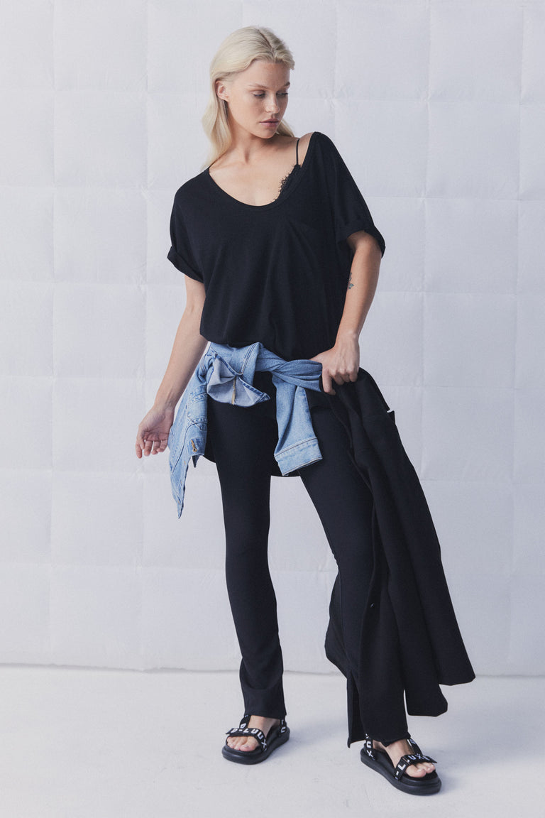 Woman wearing a relaxed black top with lace detail, black slim pants, denim jacket tied at waist, and black sandals.