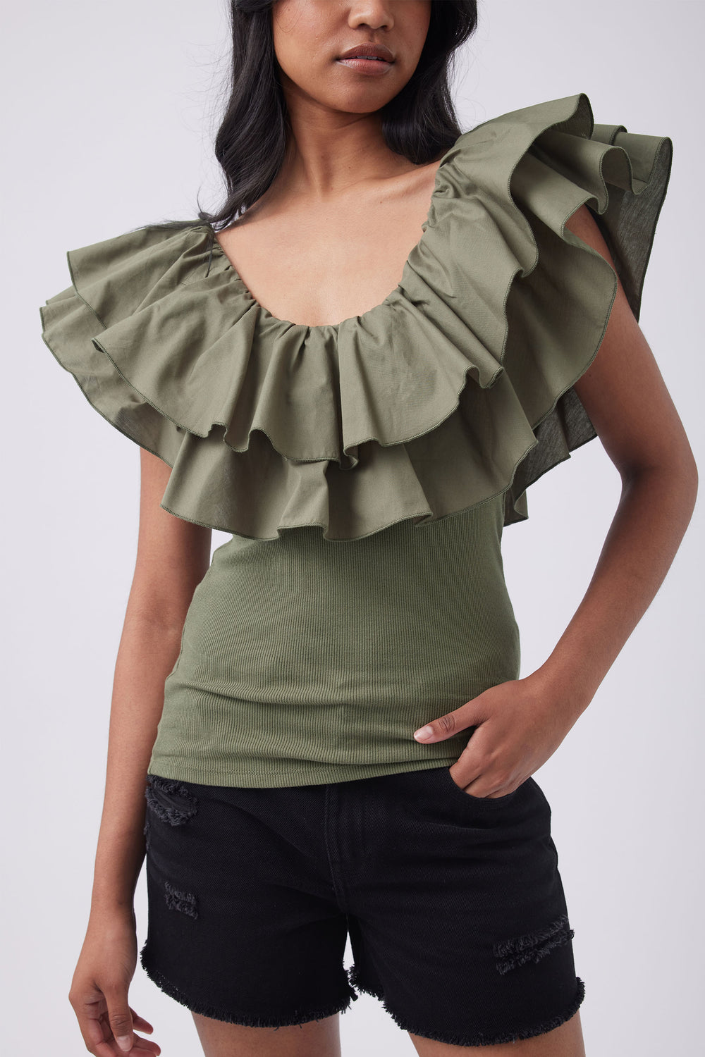 Woman wearing an olive green sleeveless top with a layered ruffle neckline and black denim shorts