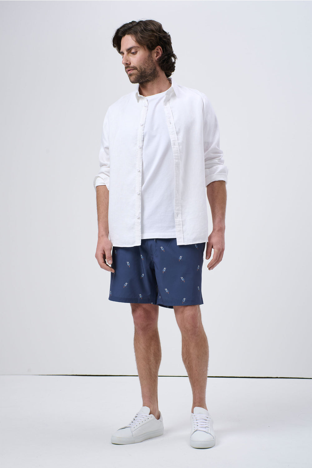 Man wearing navy swim shorts with small light print, paired with white T-shirt and open white button-up shirt, casual style