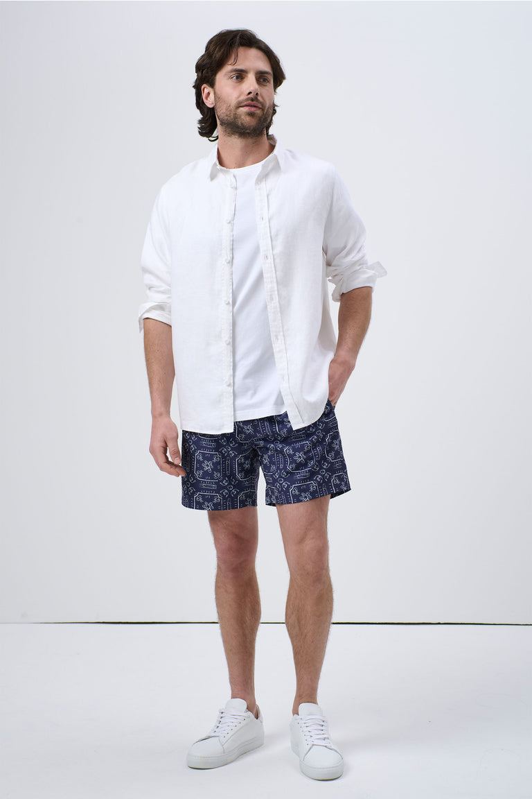 Man wearing navy patterned swim shorts paired with a casual white open shirt and white sneakers, relaxed pose