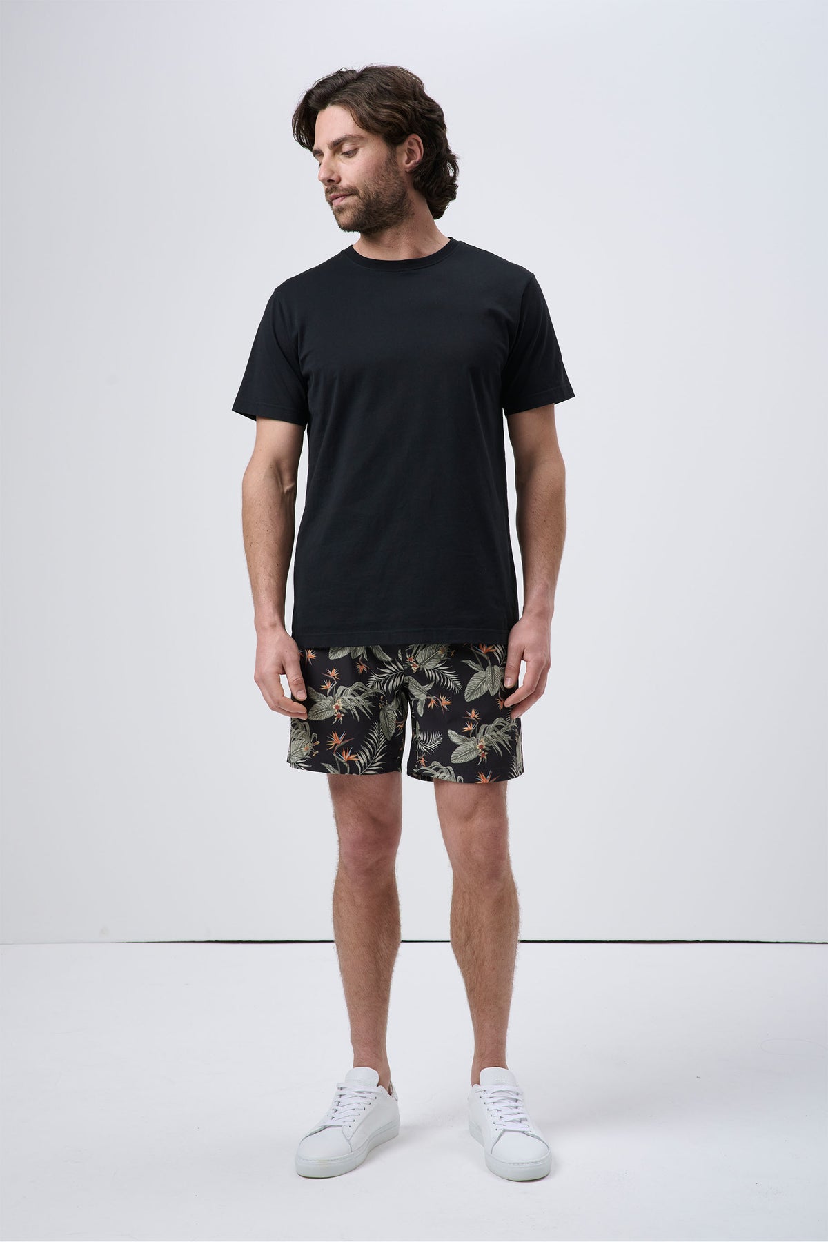 Man wearing black crew neck t-shirt paired with tropical print swim shorts and white sneakers, casual relaxed look