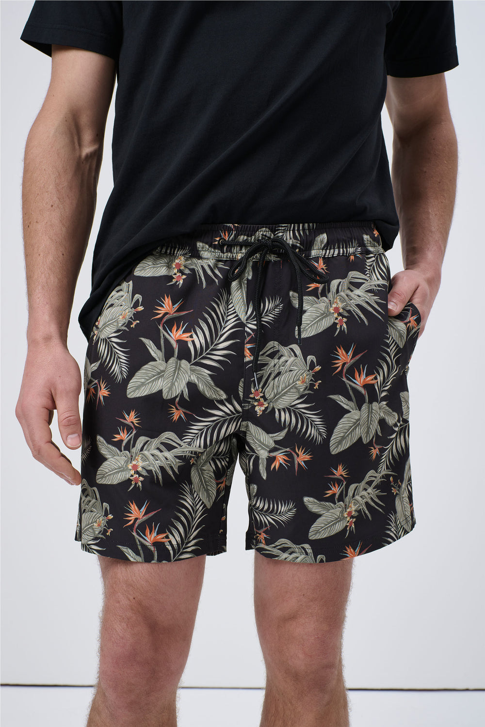 Men’s swim shorts with a tropical floral print in muted greens and orange on black, styled with a black tee.