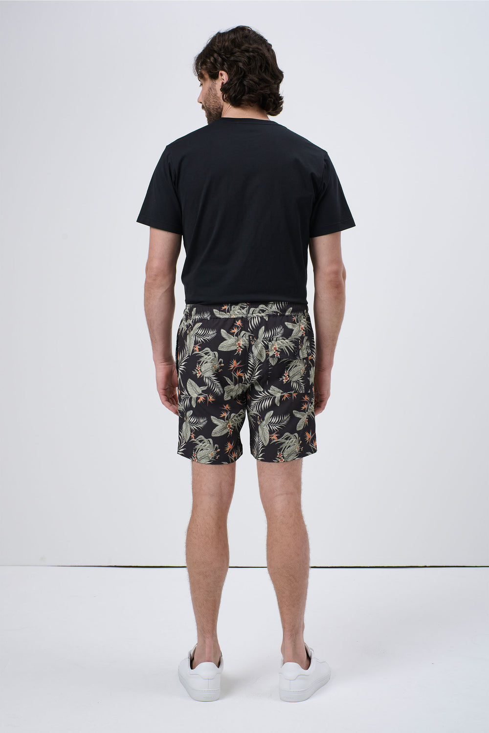 Man wearing black tropical print swim shorts paired with a plain black T-shirt and white sneakers, standing back view