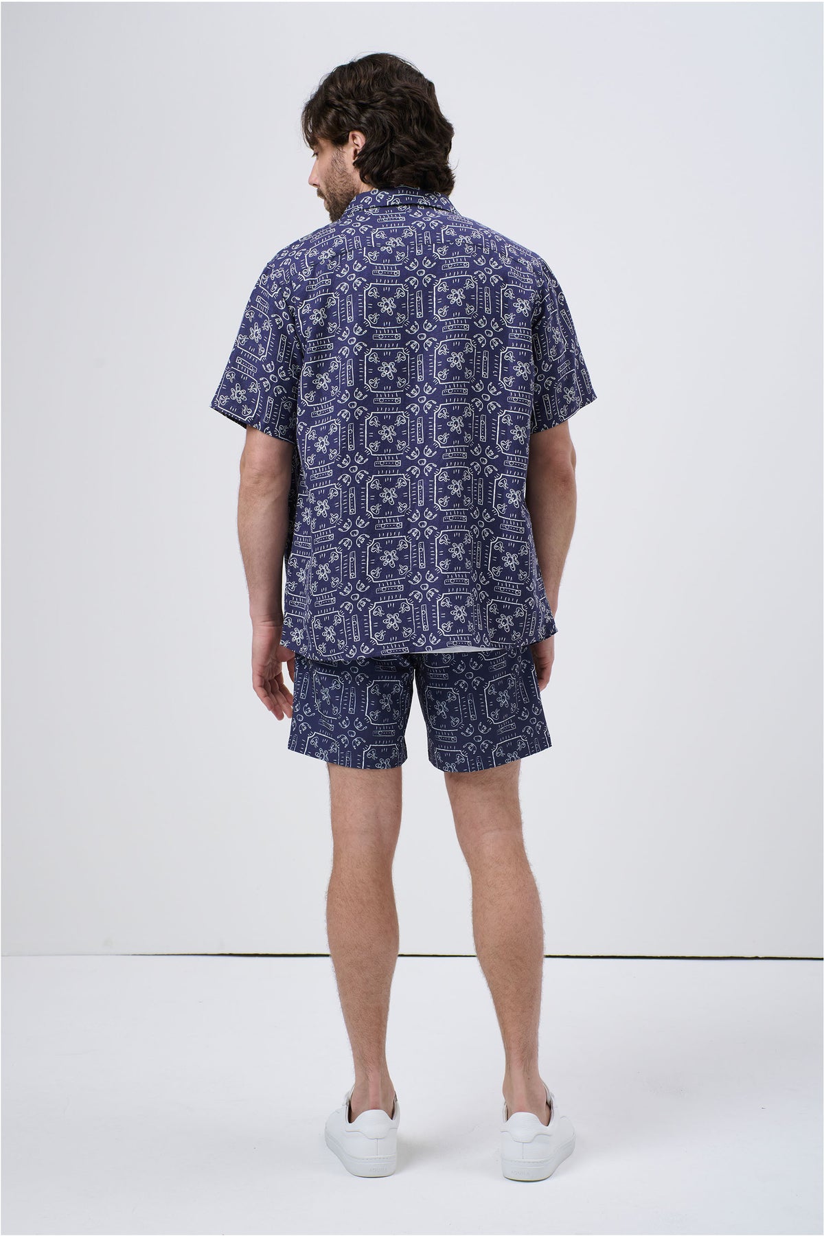 Man wearing a navy patterned short sleeve Cuban shirt and matching shorts, styled casually with white sneakers, back view