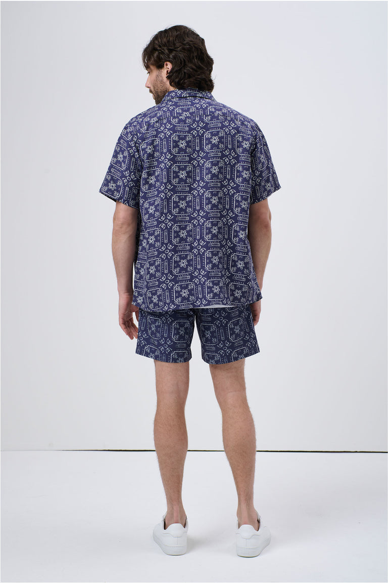 Man wearing a navy patterned short sleeve Cuban shirt and matching shorts, styled casually with white sneakers, back view