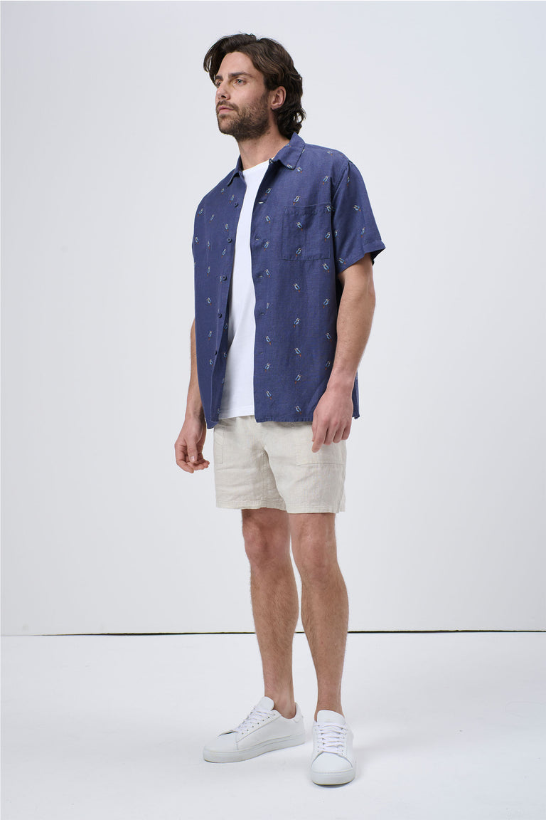Man wearing a navy short-sleeve Cuban shirt with subtle print over a white tee and beige shorts, styled casually with white sneakers