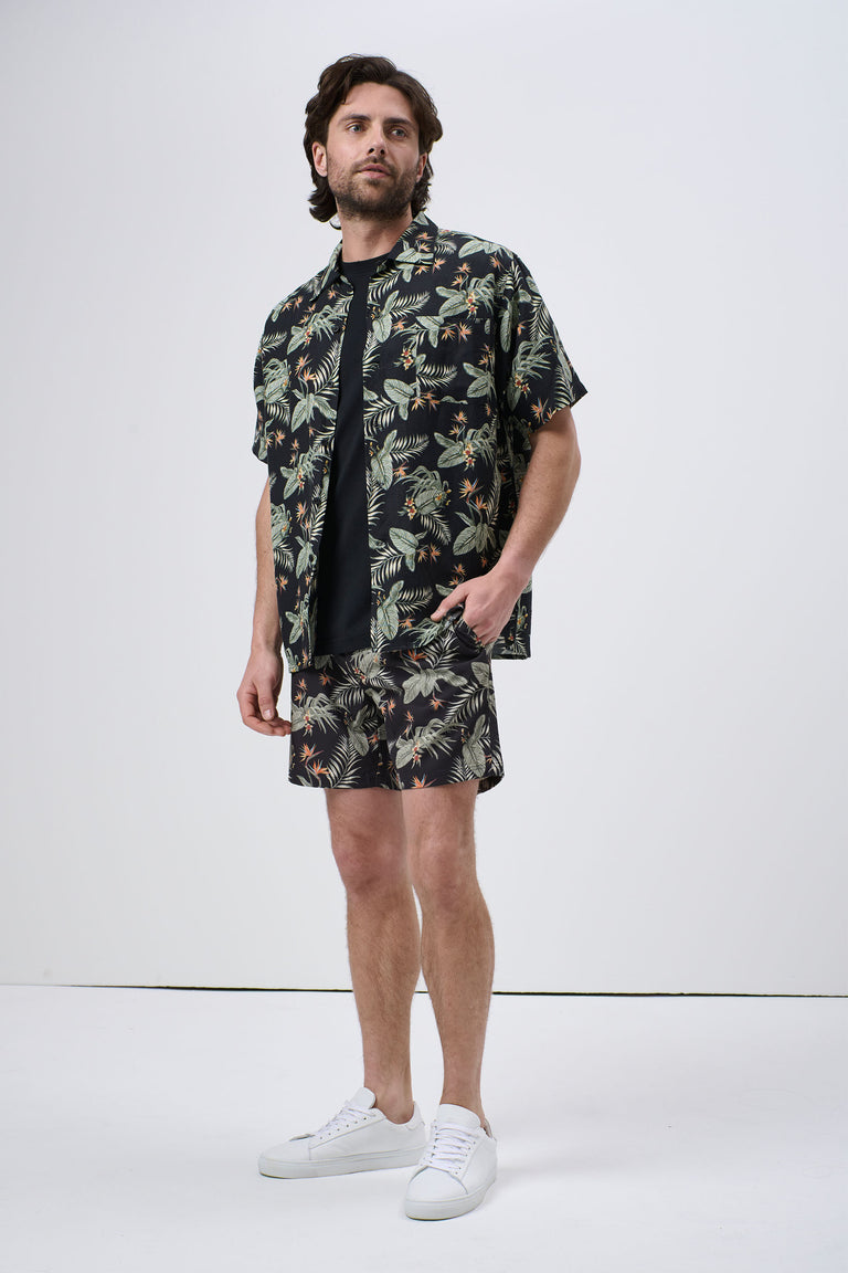 Man wearing a black Cuban shirt with green and beige tropical leaf print, paired with matching shorts and white sneakers