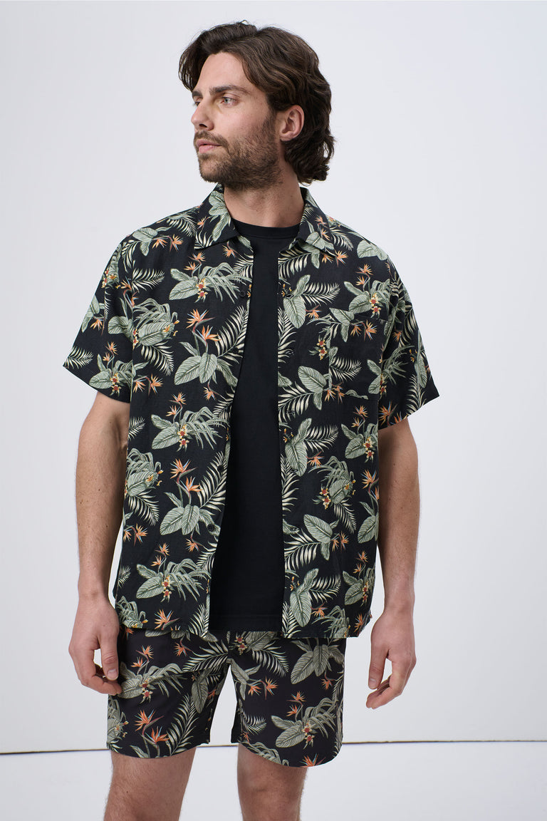 Man wearing a dark tropical print short-sleeve shirt layered over a black tee, paired with matching shorts, casual style
