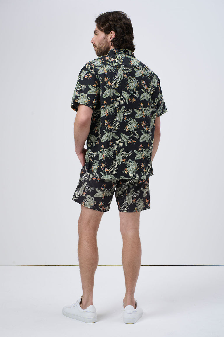 Man wearing a black short-sleeve Cuban shirt with green tropical leaf print and matching shorts, standing with hands in pockets