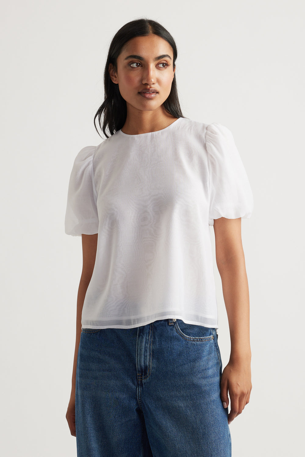 Woman wearing a white puff sleeve top with round neckline, paired with relaxed blue denim jeans against a plain background