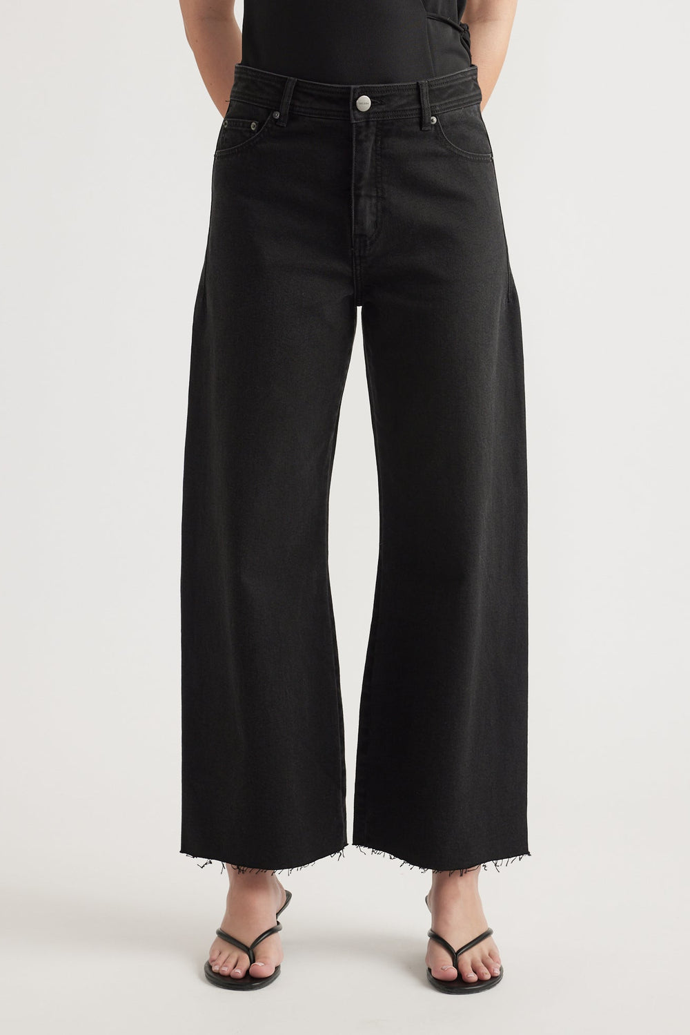 High rise black wide-leg cropped jeans with raw hem styled with black strappy sandals on a neutral background