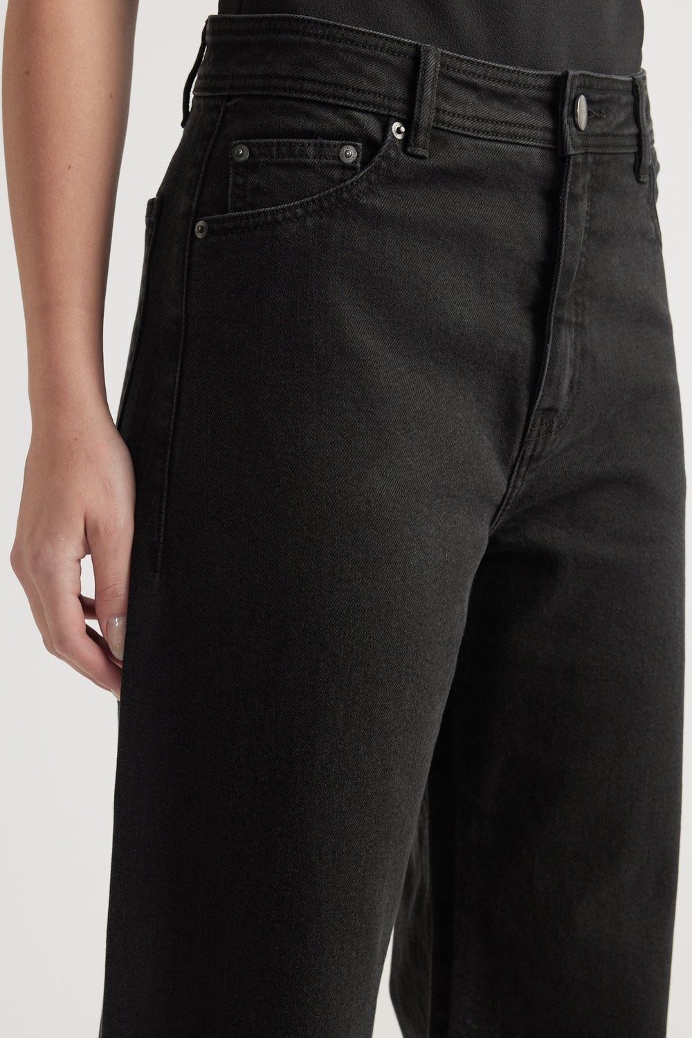 Close-up of high-rise black denim jeans with wide leg and subtle front pocket detailing on model.