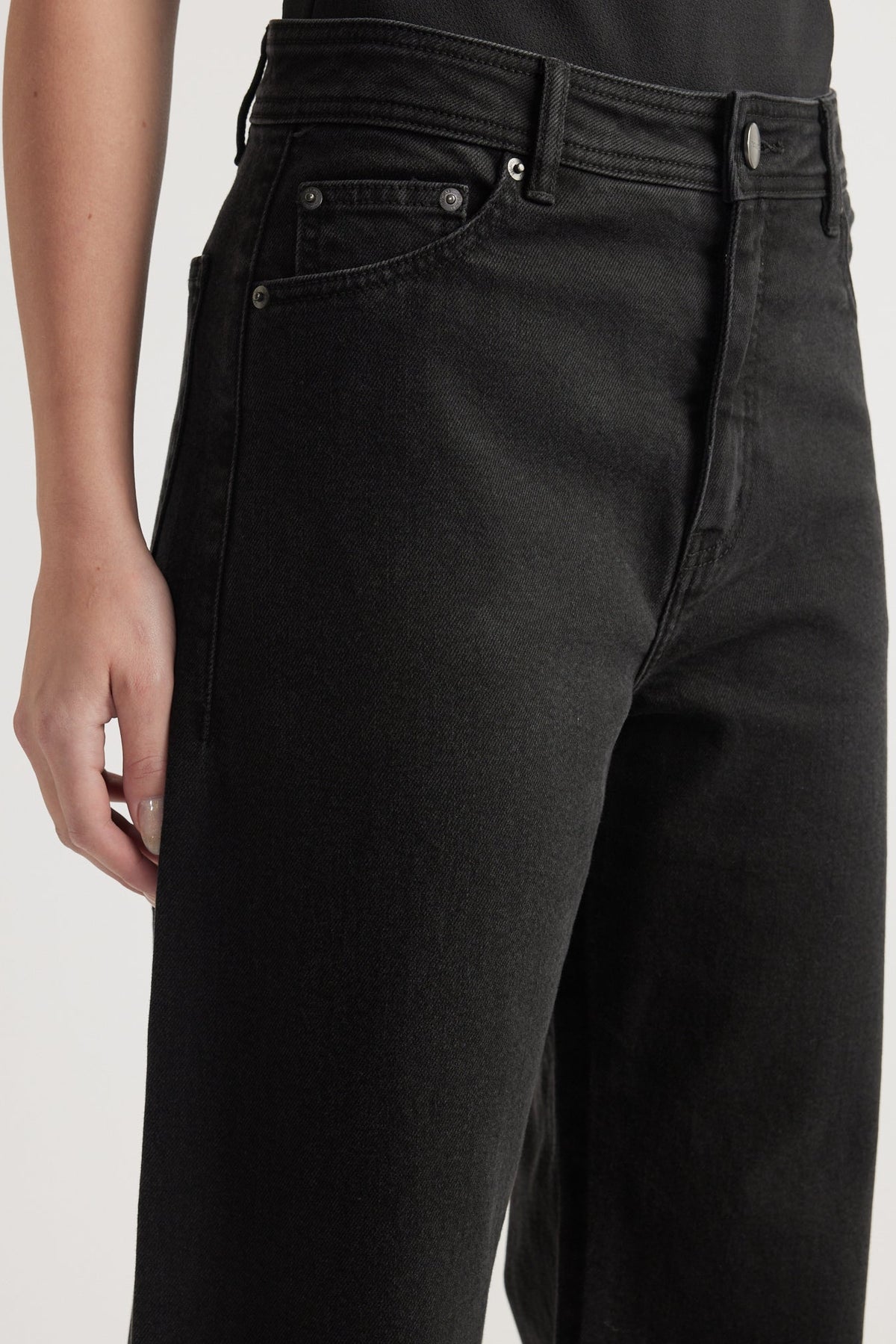 Close-up of high-rise black denim jeans with wide leg and subtle front pocket detailing on model.