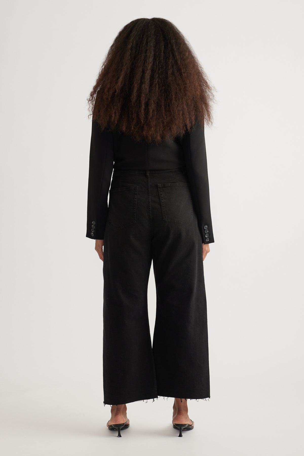 Back view of woman wearing high rise black wide leg jeans with cropped raw hems, black top, and heeled sandals