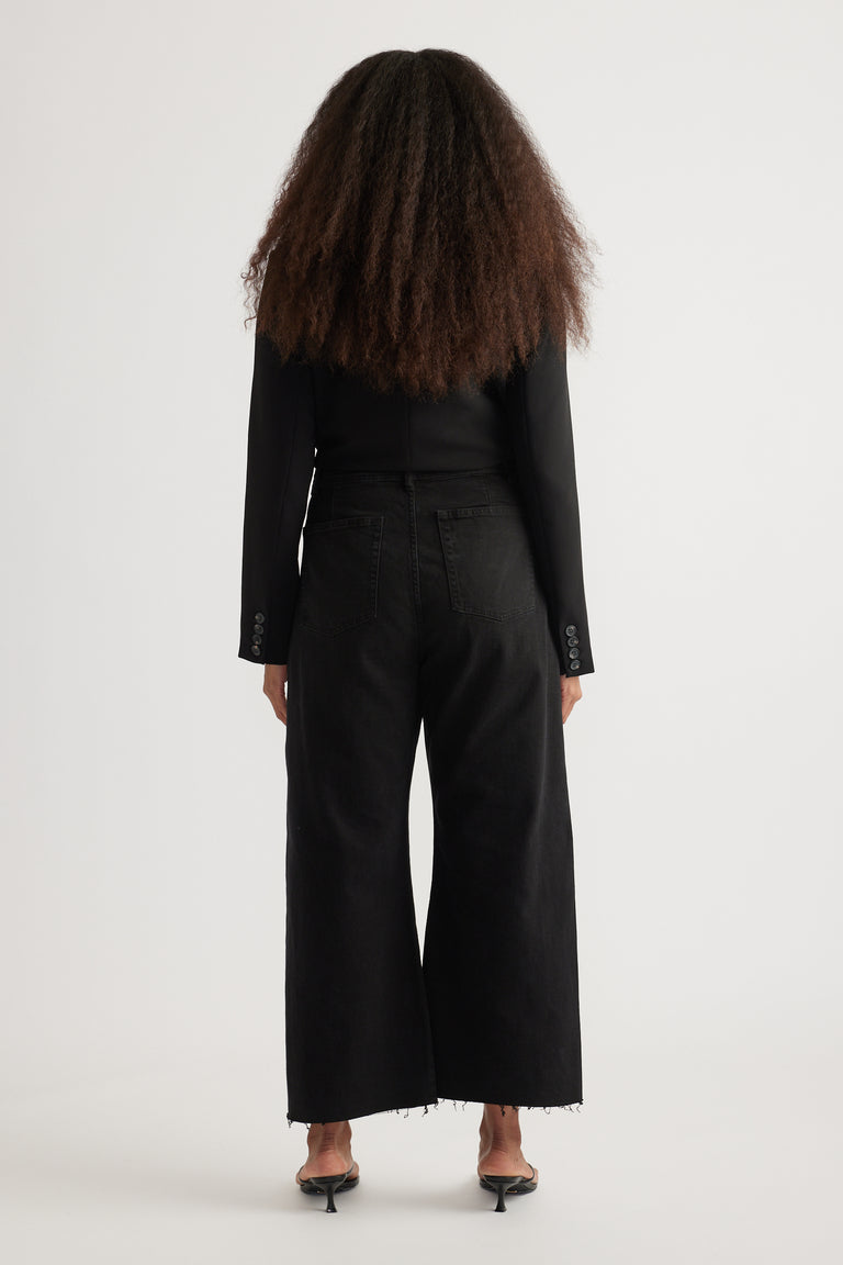 Back view of woman wearing high rise black wide leg jeans with cropped raw hems, black top, and heeled sandals