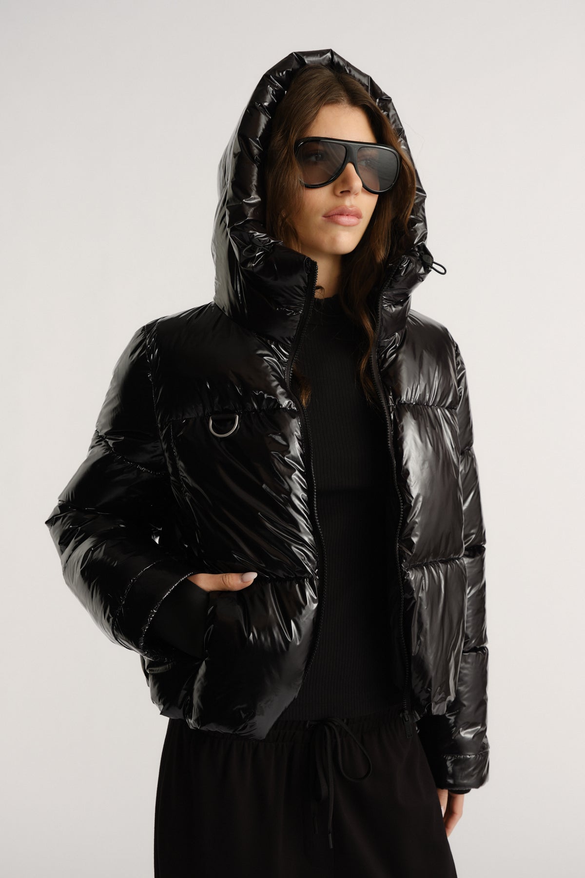 Woman wearing a glossy black puffer jacket with a hood, styled with oversized sunglasses and black knitwear underneath.