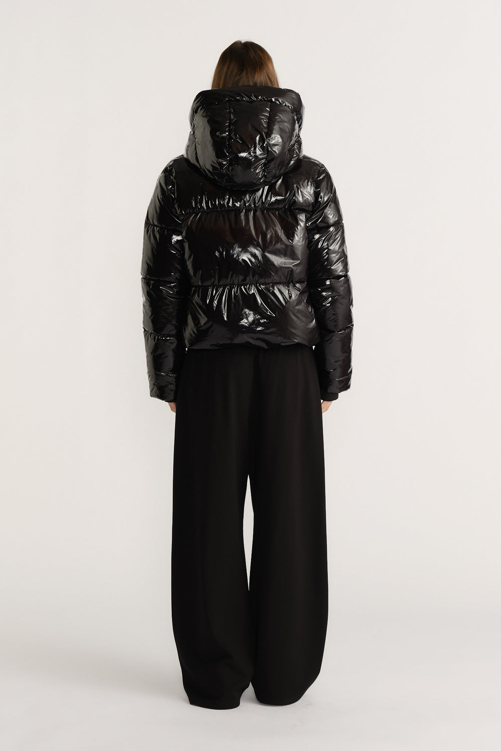 Woman wearing a shiny black puffer jacket with a large hood paired with wide-leg black pants, back view.
