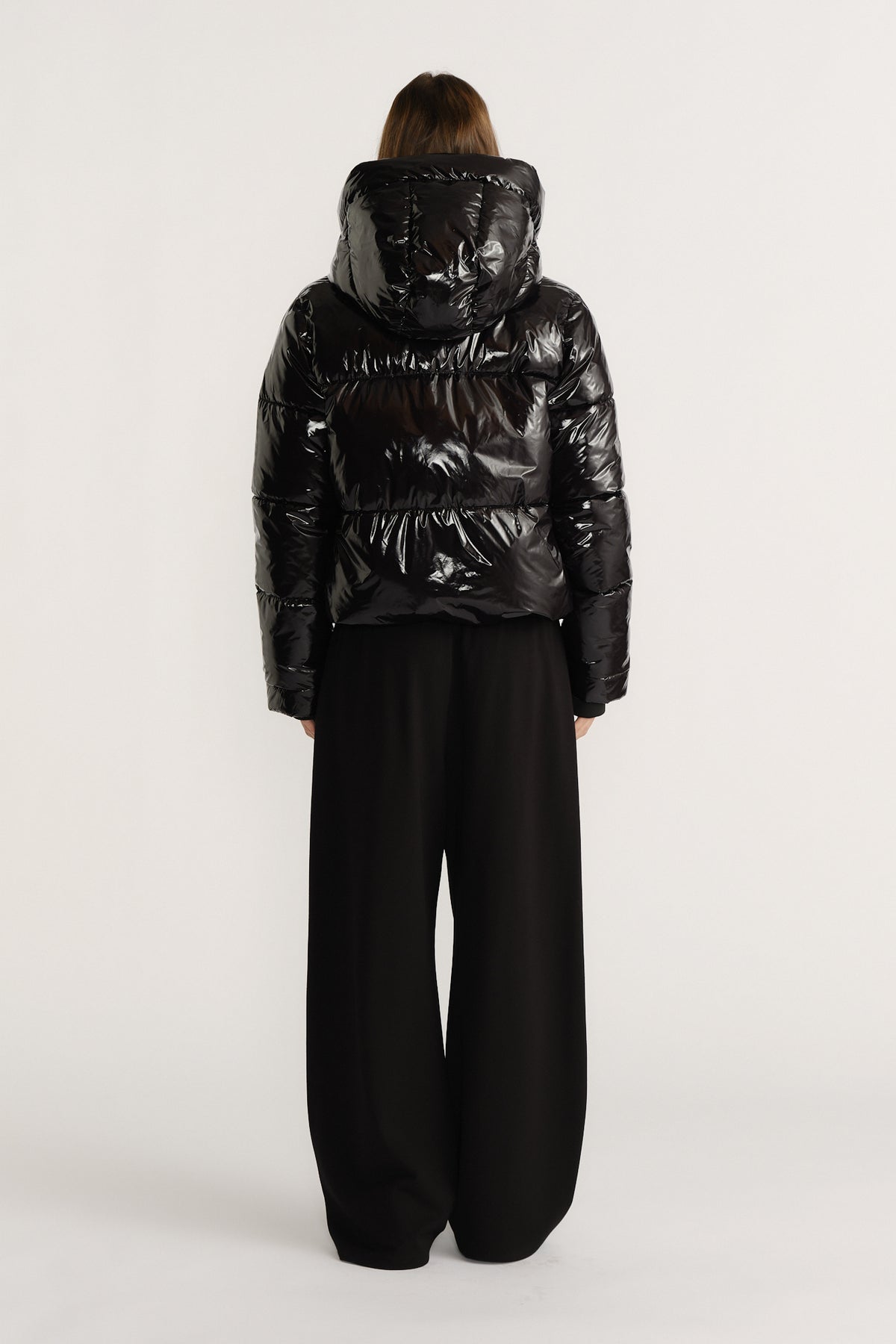 Woman wearing a shiny black puffer jacket with a large hood paired with wide-leg black pants, back view.