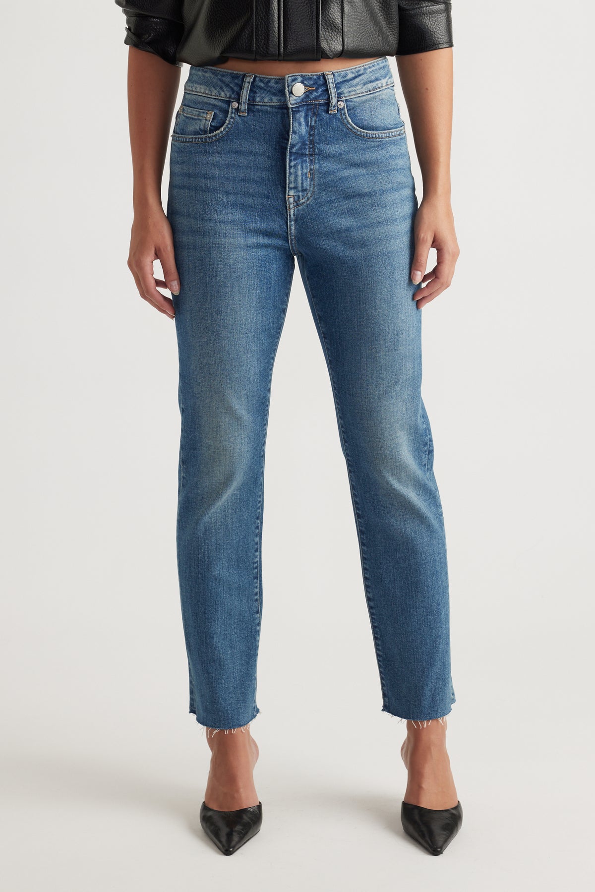 Front view of mid-blue straight-leg high-rise jeans with subtle fading and raw hem, styled with black pointed flats.