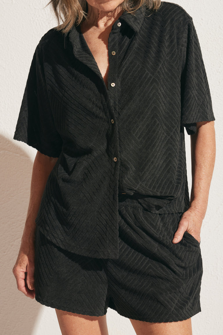 Woman wearing a relaxed black terry shirt with subtle textured chevron pattern and matching shorts, hands in pockets