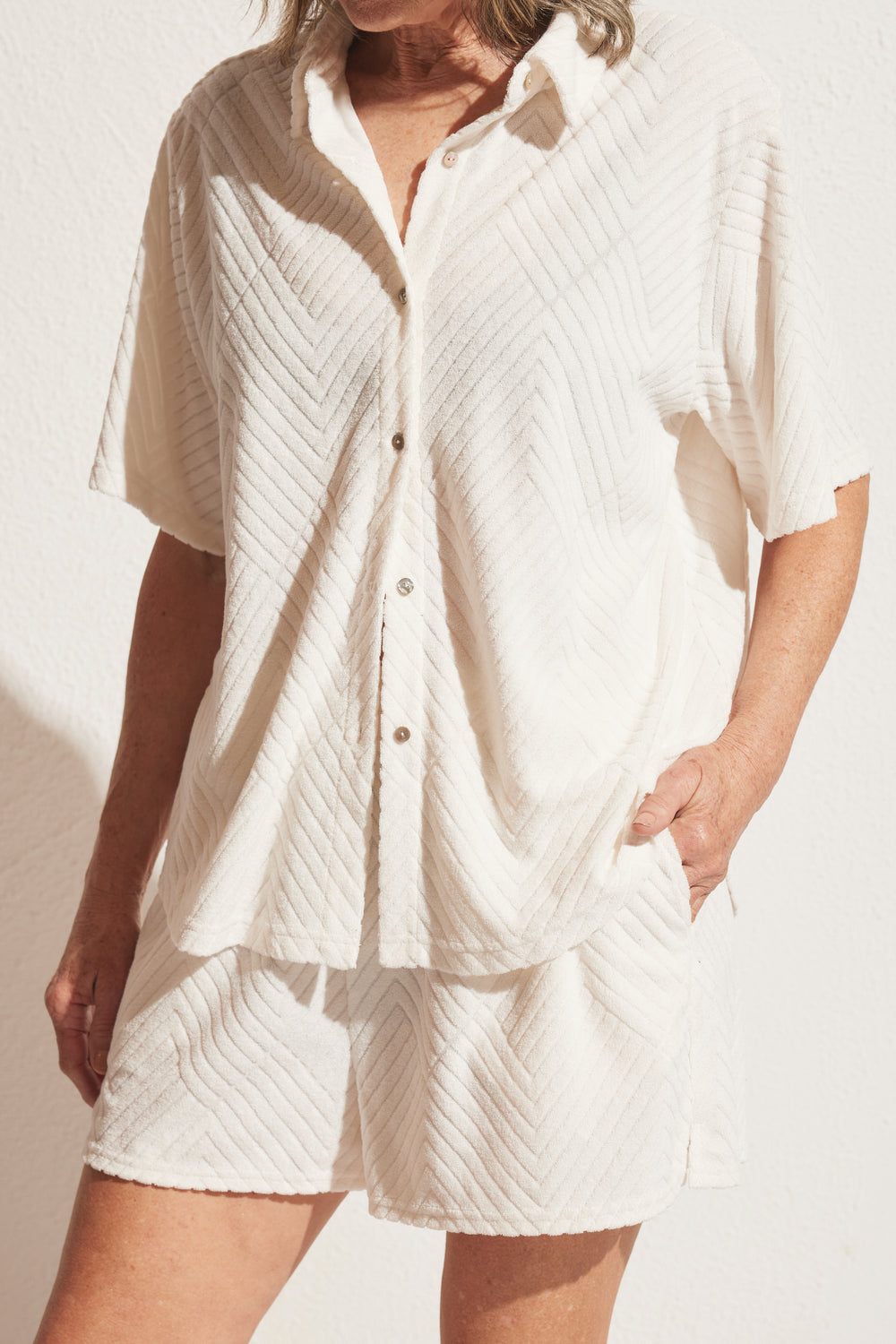 Woman wearing textured cream short-sleeve button-up top and matching relaxed shorts with hand in pocket