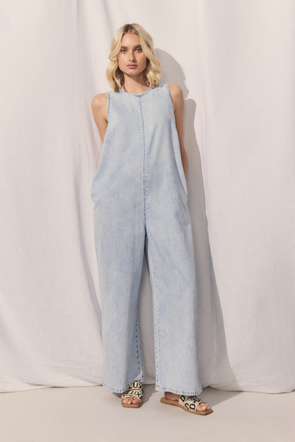 Portrait of woman wearing sleeveless D-Luxe Palazzo Denim Jumpsuit with straight leg and side pockets, gallery image 1.