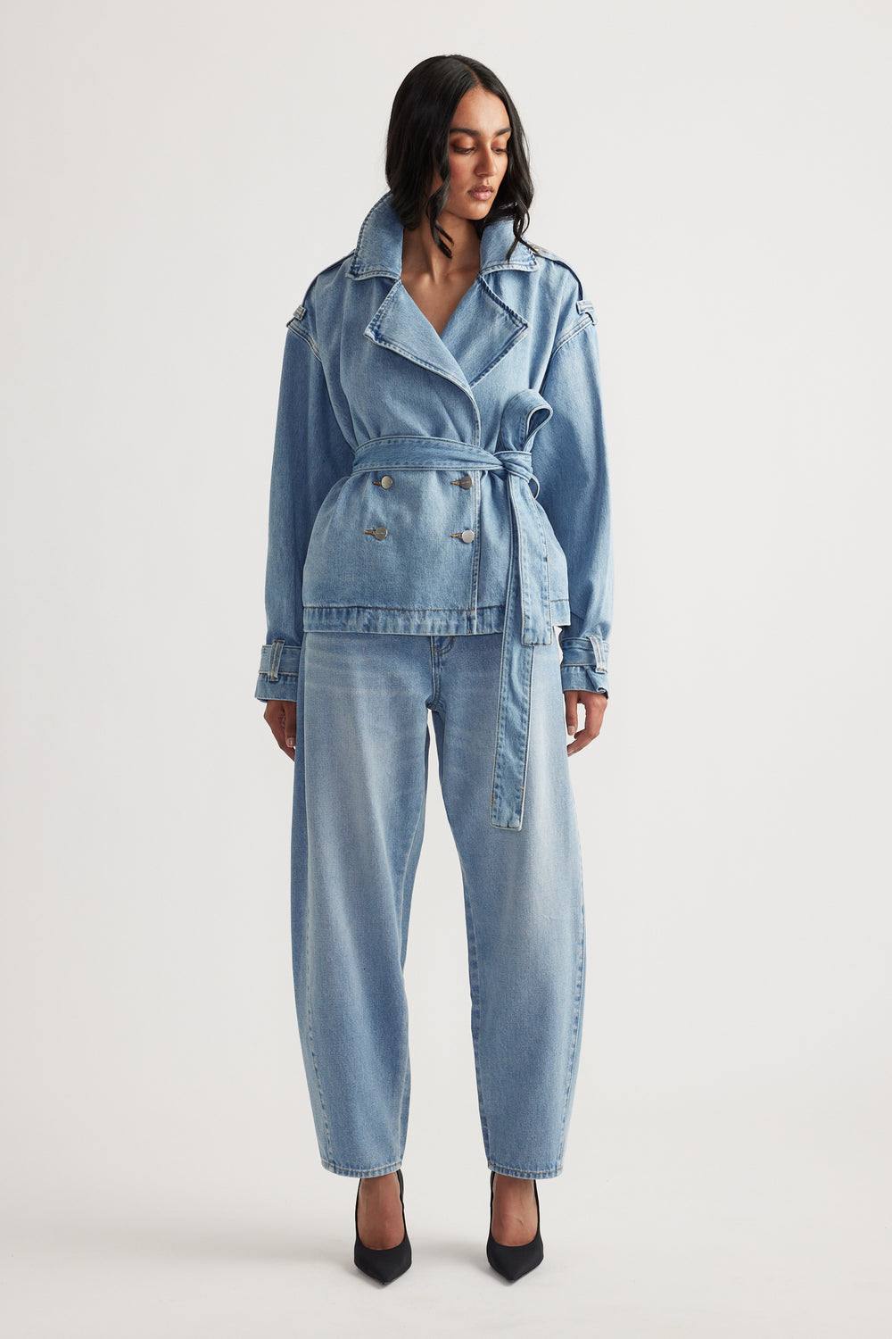 Woman wearing light blue belted denim jacket paired with relaxed fit barrel leg jeans and black pointed heels
